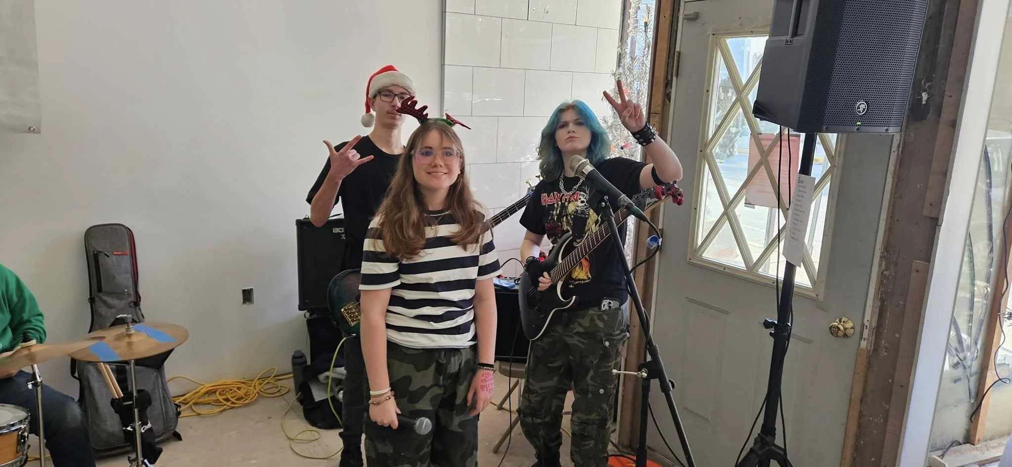 Three young women with music instruments in a room decorated for a holiday event. Two are holding microphones, one has a guitar, and they are making peace signs with their hands. One is wearing a Santa hat with reindeer antlers, and another has color