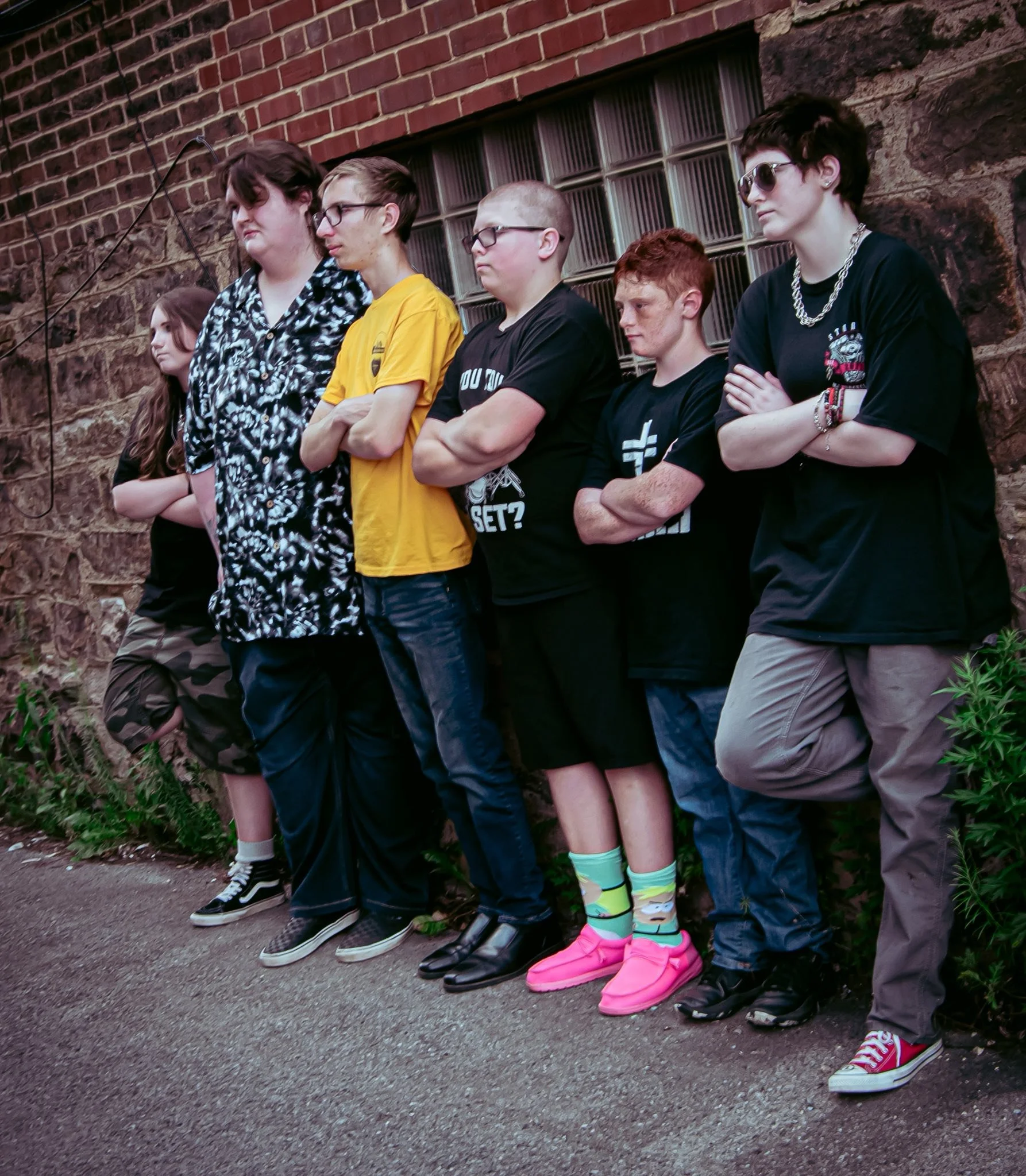 Six teenagers stand with arms crossed against a brick wall, looking serious and defiant.