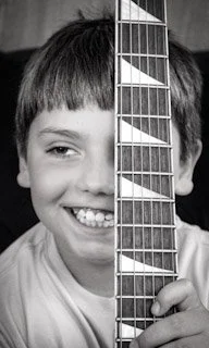 A smiling boy holding a guitar vertically covering part of his face