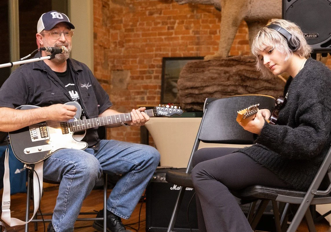 A man and a woman playing electric guitars in a music studio with brick walls, with musical equipment around them.