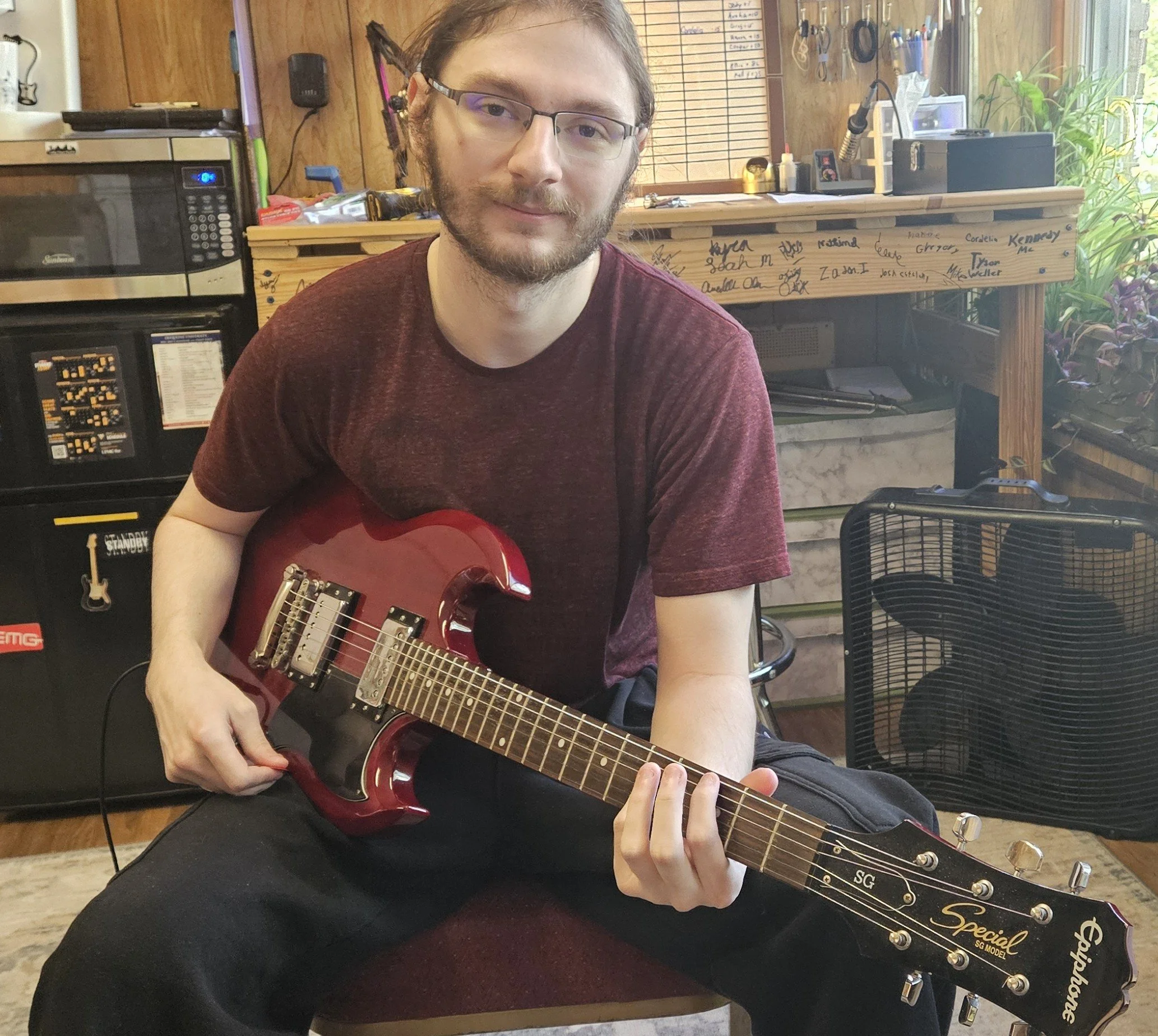 A young man with glasses and a beard sitting on a stool, holding a red electric guitar, in a wood-paneled room with appliances, tools, and handwritten notes on a shelf behind him.