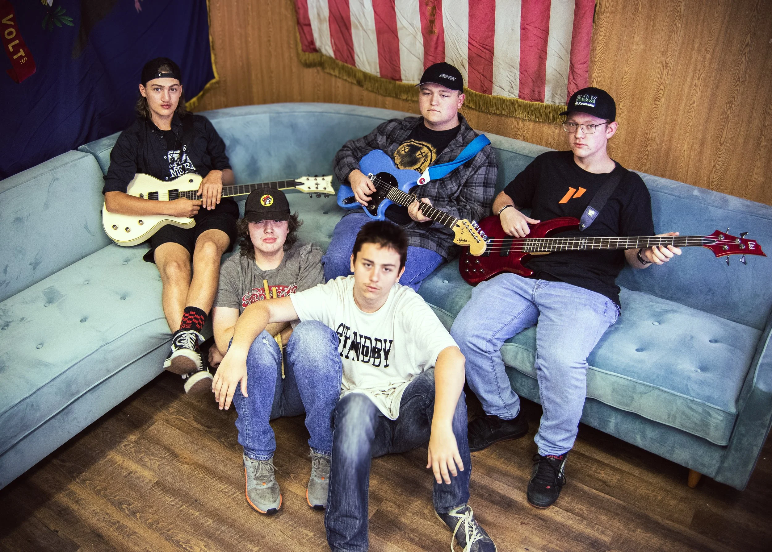 A group of five teenage friends sitting and posing with guitars on a light blue sofa in a room with wooden floors and wood-paneled walls, with patriotic flags hanging on the wall behind them.