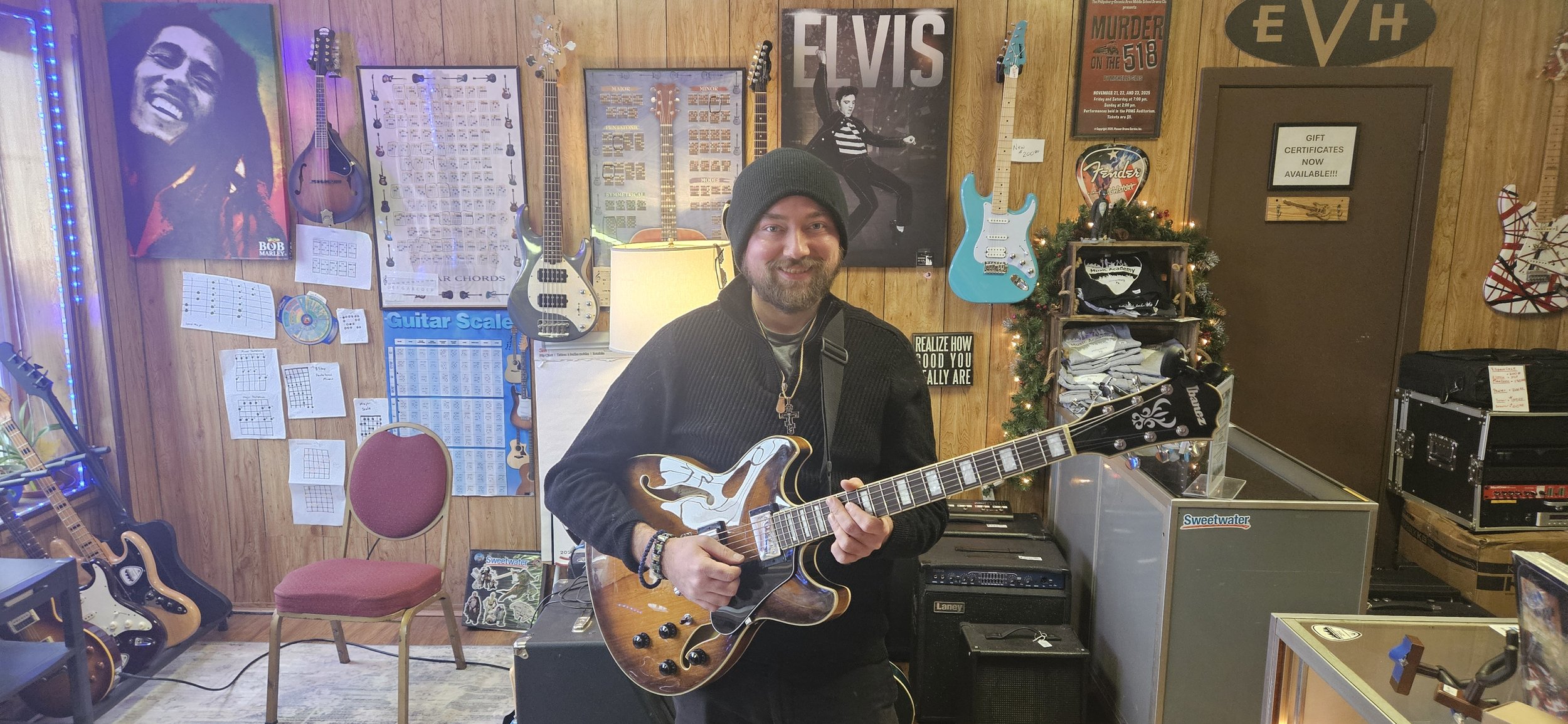 A man with a black beanie, black sweater, and a beard holding an electric guitar inside a music store decorated with guitars on the wall, posters, and music charts. The background features poster art including Elvis Presley and Bob Marley, and there are more guitars and music equipment around.
