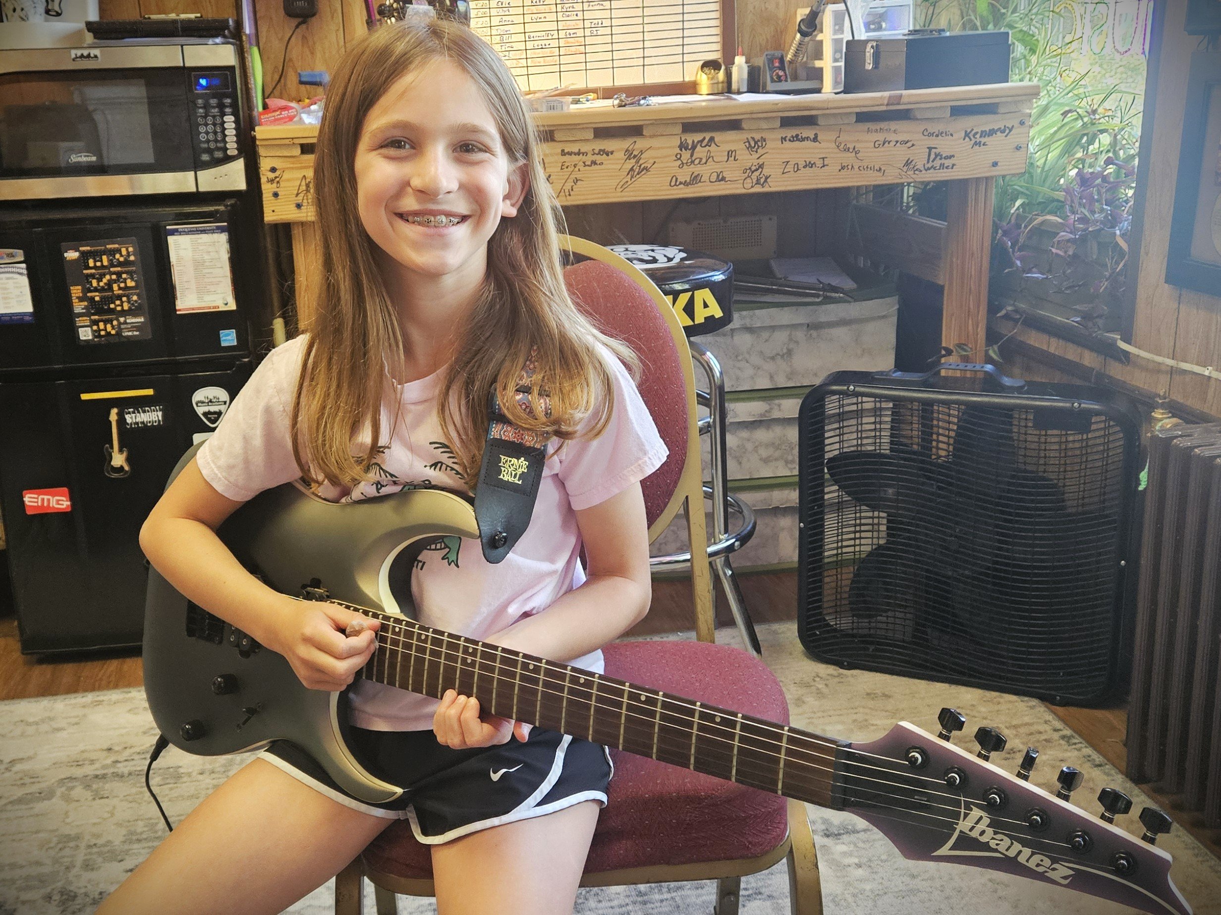 A young girl sitting on a red chair in a room, holding a black electric guitar, smiling at the camera. The room has a wooden wall, a black fan, a microwave, a small table, and some plants on the windowsill.