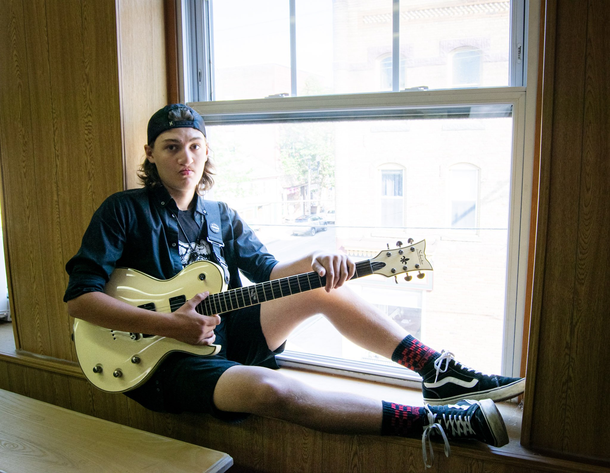 A young person with long hair, wearing a black cap and dark clothing, sitting on a windowsill while holding a white electric guitar.