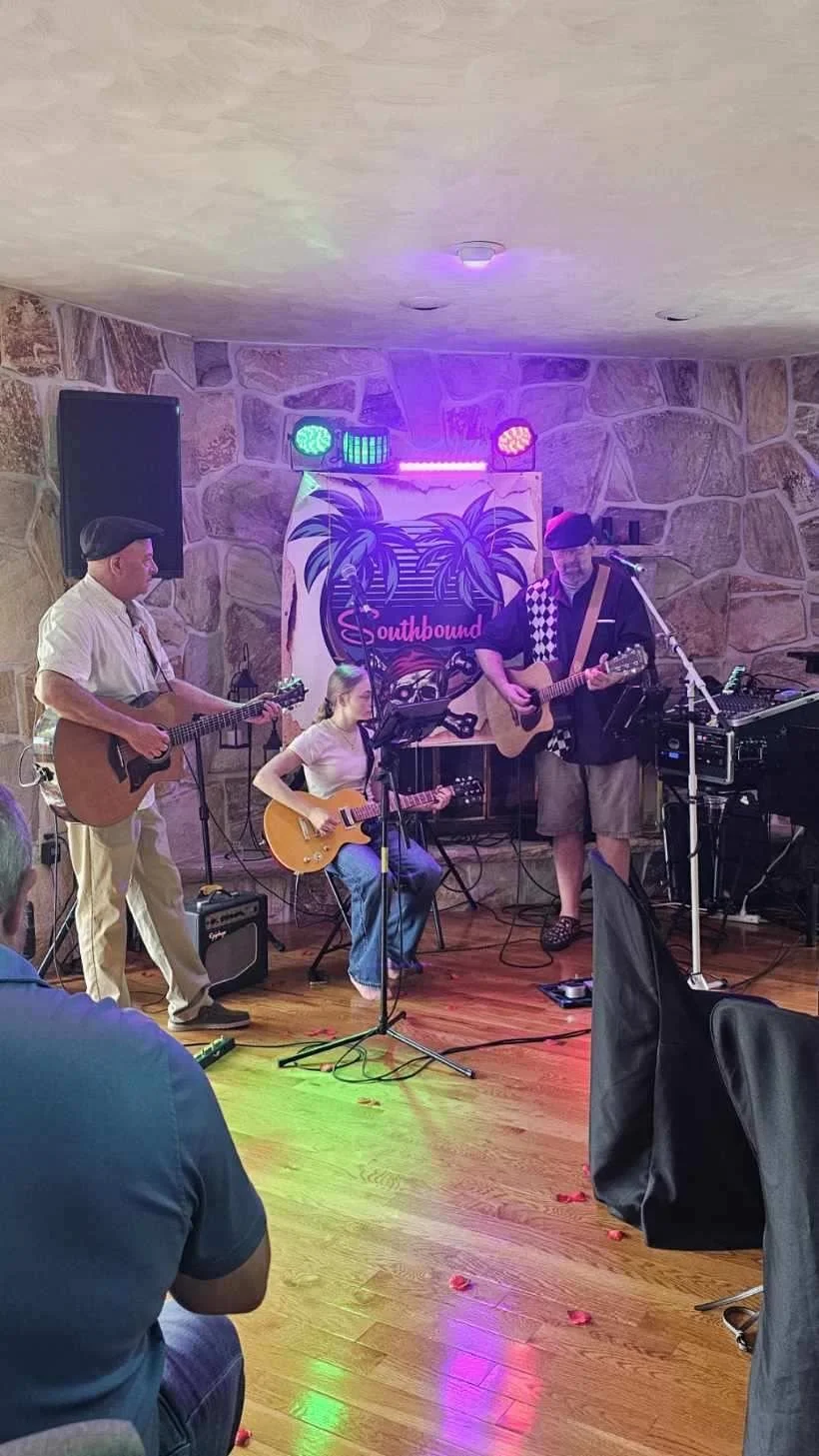 A band of three musicians performing with guitars on a small indoor stage decorated with a Miami-themed backdrop, colorful lights, and a stone wall, with audience members watching.
