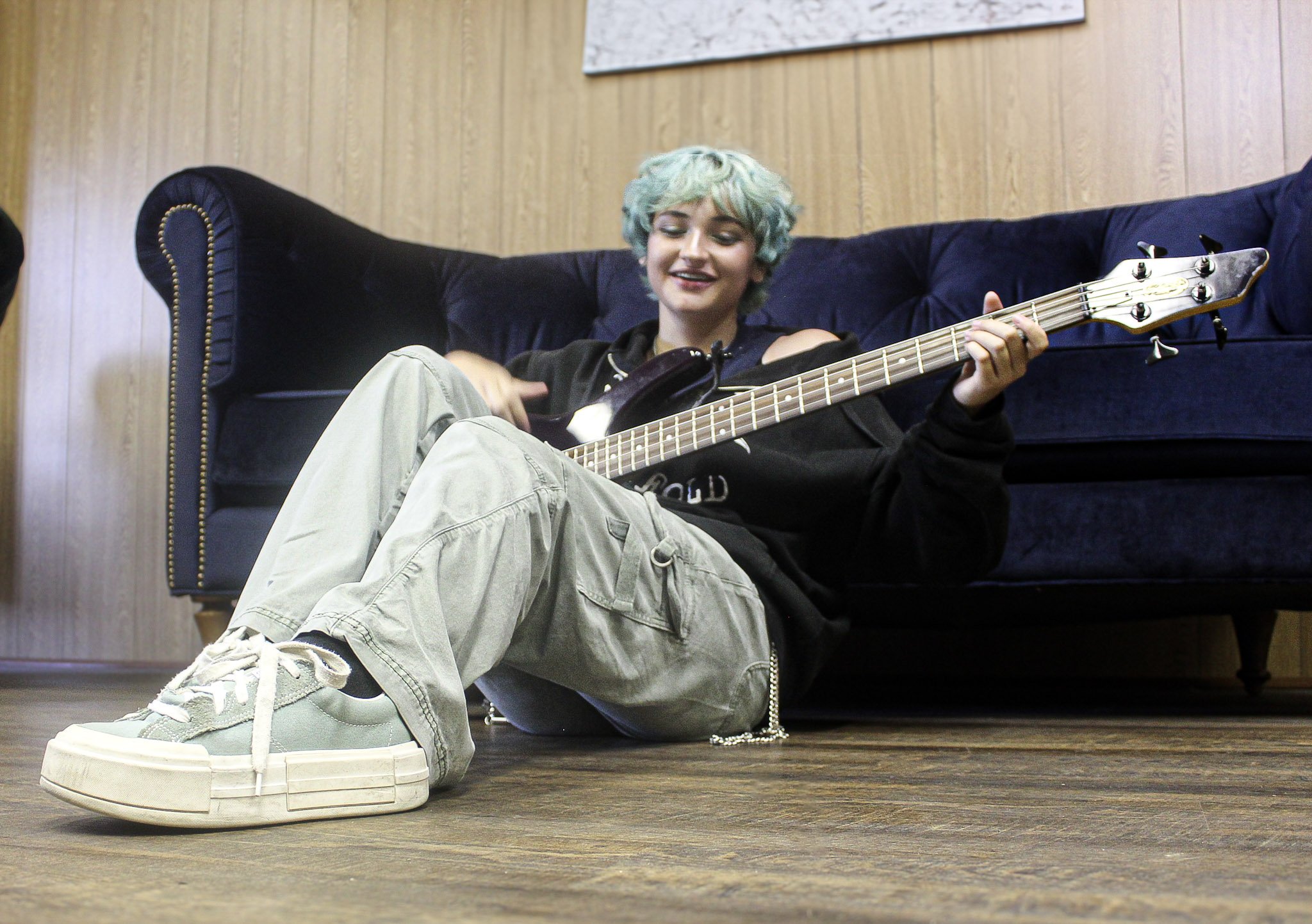 A young person with short, curly, light blue hair sitting on the floor in front of a blue velvet couch, playing a bass guitar, smiling.