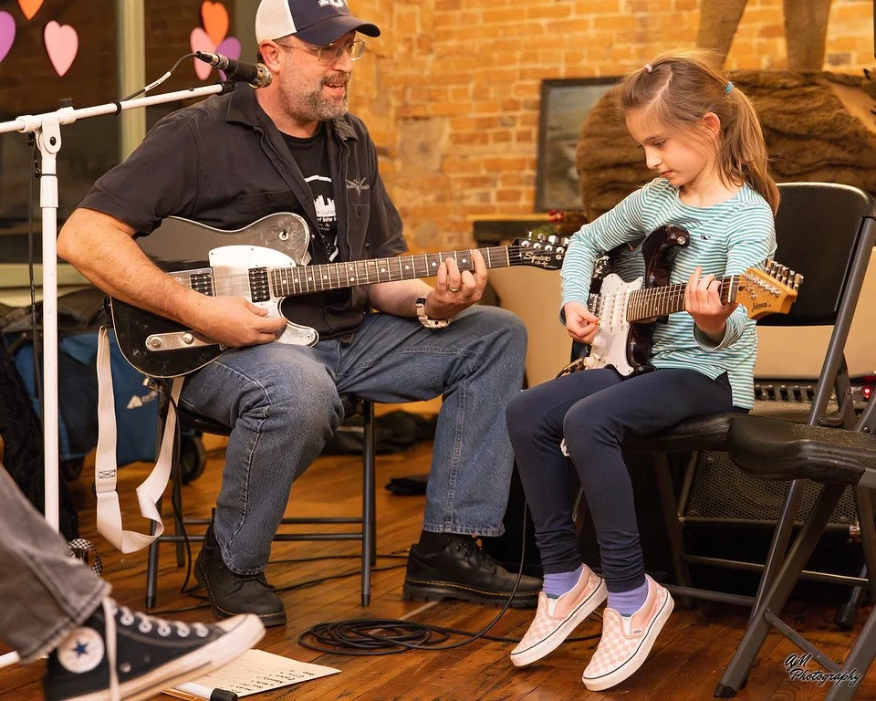 A man and a young girl are playing electric guitars together, seated in a room with brick walls, wood flooring, and musical equipment. The man is wearing a black cap, glasses, and a black shirt, while the girl has a striped shirt and checkered Vans s