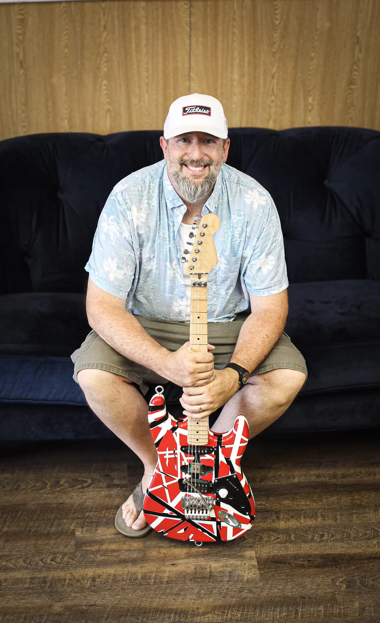 A man with a beard and wearing a baseball cap sitting on a black couch, holding an electric guitar with a red, black, and white abstract design, in a room with wood-paneled walls and a wooden floor.