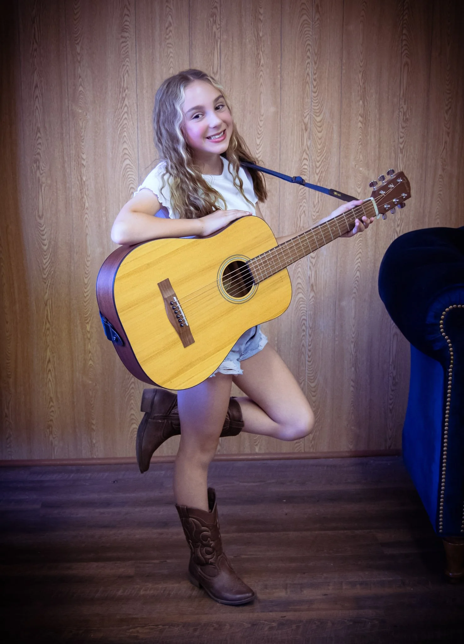 A young girl with long curly hair, wearing a white t-shirt, shorts, and cowboy boots, standing on one leg while playing an acoustic guitar. She is smiling and posing indoors against a wood-paneled wall.