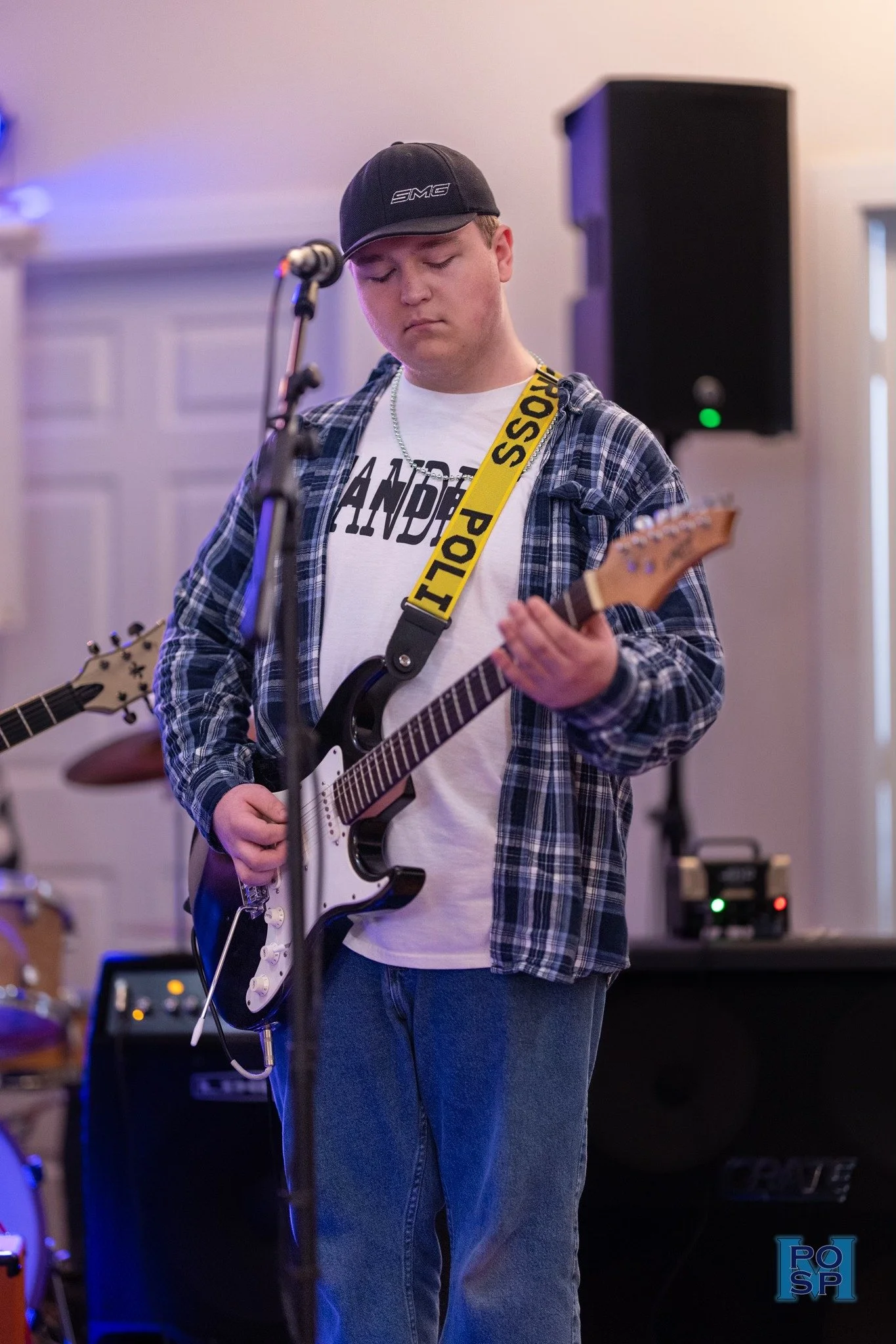 Young man playing electric guitar during a live performance, wearing a black cap, plaid shirt, and a white T-shirt, with a yellow 'POLICE' strap around his neck.