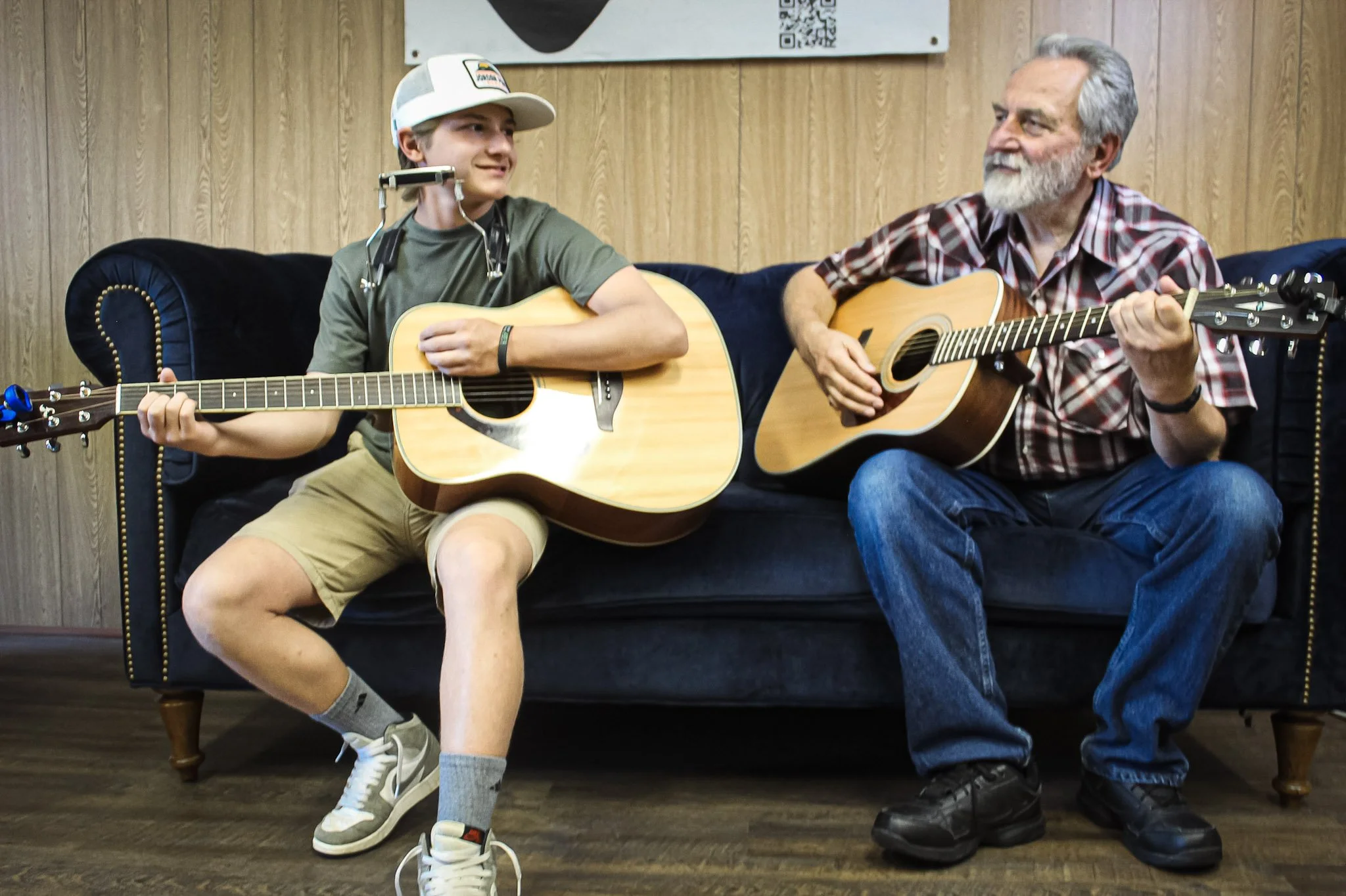 A boy and an older man sit on a black couch playing acoustic guitars, looking at each other and smiling while playing music in a room with wood-paneled walls.