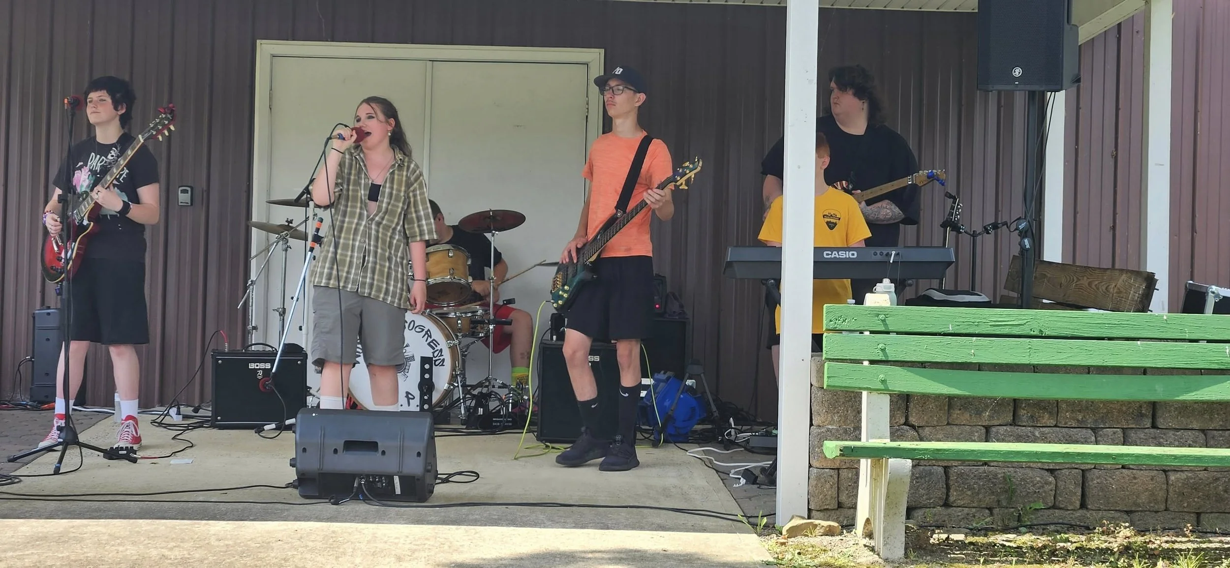 A band performing on an outdoor stage with five members playing various instruments: two guitarists, a bassist, a keyboardist, and a drummer, with a green bench in the foreground.