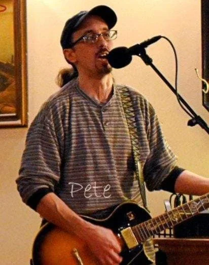 Man singing into a microphone while playing an electric guitar, wearing glasses, a cap, and a striped shirt.