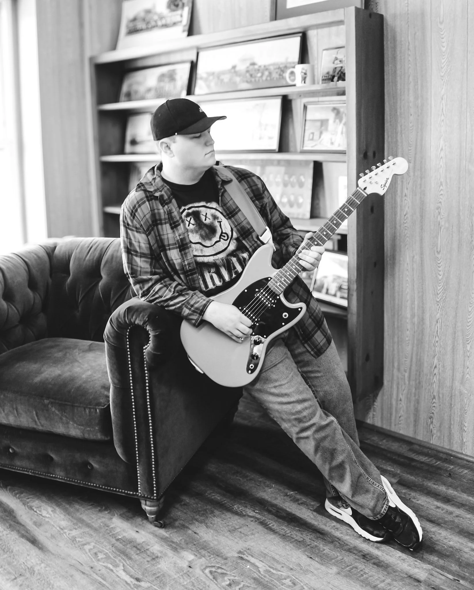 A young man wearing a baseball cap, plaid shirt, and graphic t-shirt is sitting on a bench playing an electric guitar indoors.