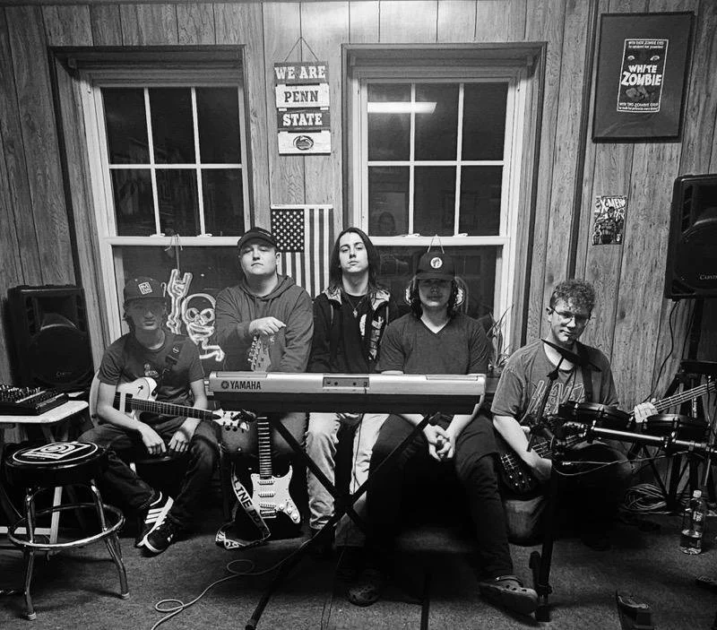 Five young people in a room with woodpanel walls, some holding musical instruments, standing and sitting behind a Yamaha keyboard, with posters and an American flag on the wall.