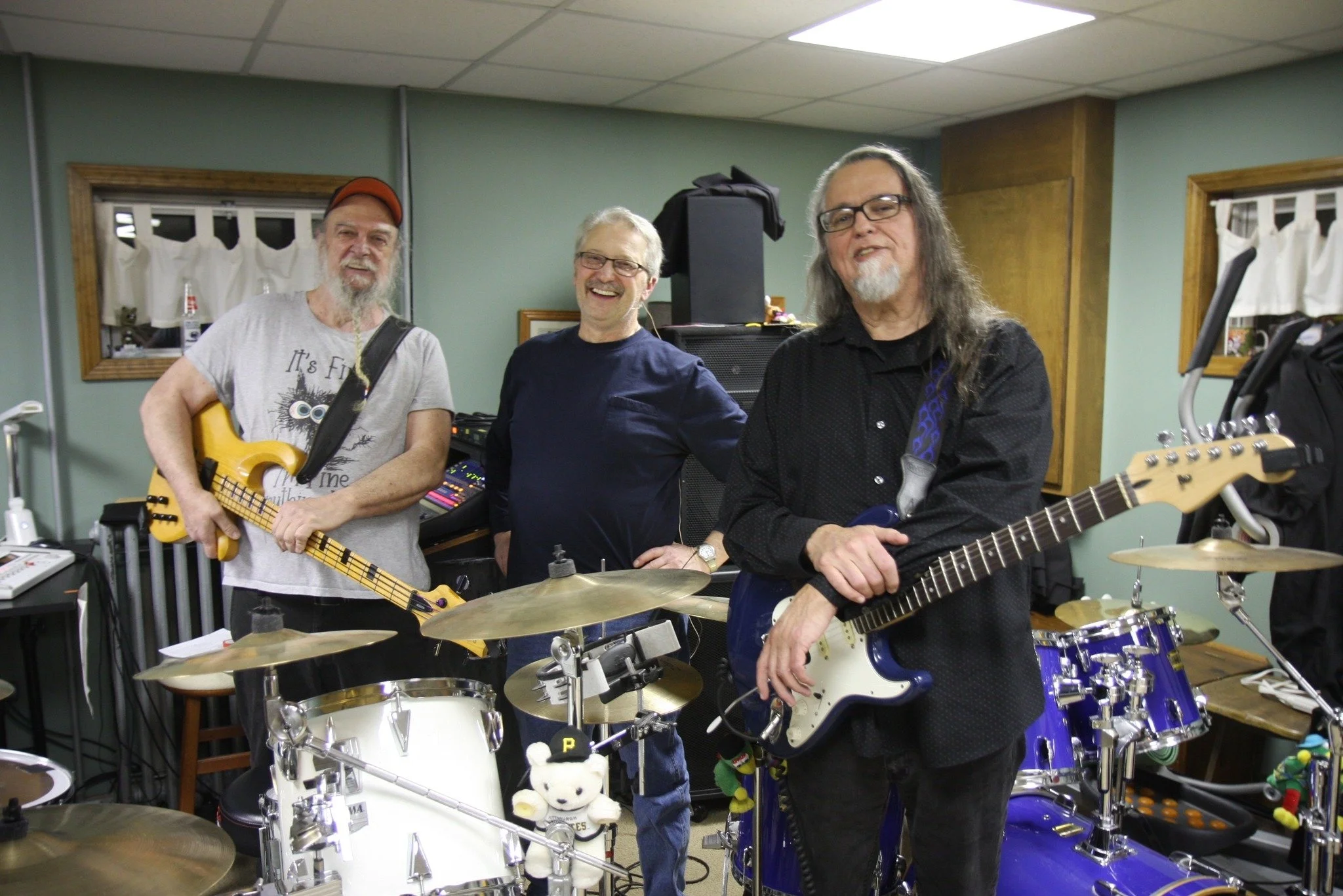 Three middle-aged men with long hair and glasses, playing musical instruments in a rehearsal room. One has a guitar, another has a bass, and the third is near drums. They are smiling and appear to be enjoying their practice.