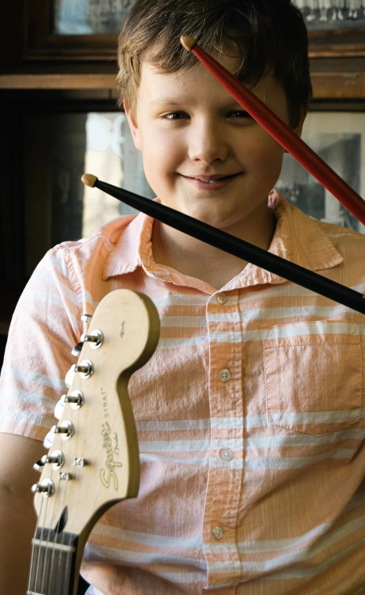 A young boy holding an electric guitar and smiling, with drumsticks crossing in front of his face, indoors near a window.