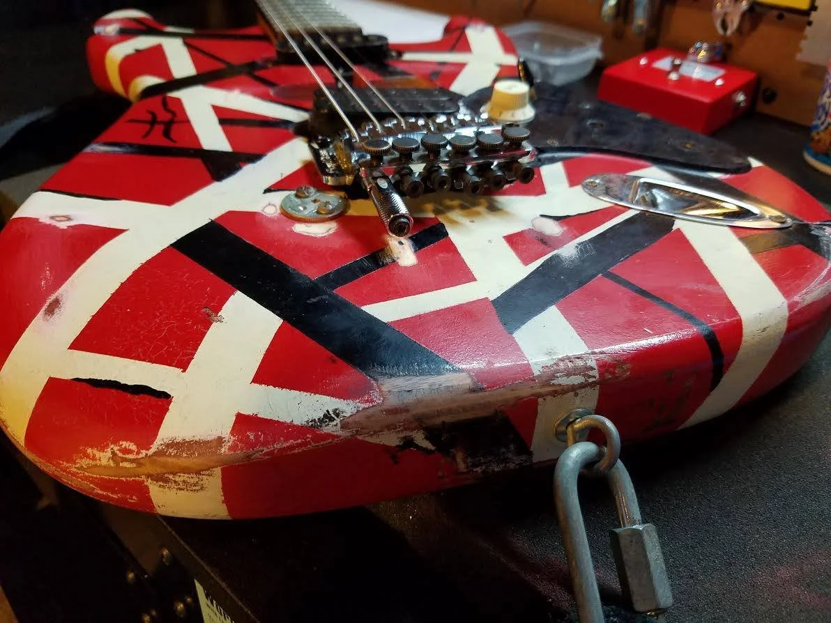 Close-up of a worn electric guitar resembling a British flag, with visible scratches and chipping paint, resting on a table with various objects in the background.