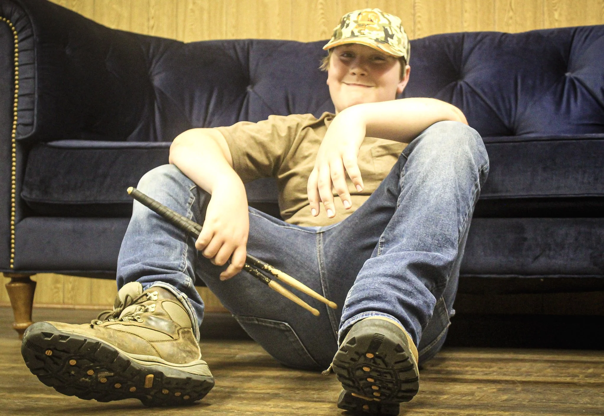 Young boy sitting on wooden floor in front of sofa, wearing camouflage cap, casual t-shirt, jeans, and hiking boots, holding drumsticks, smiling.