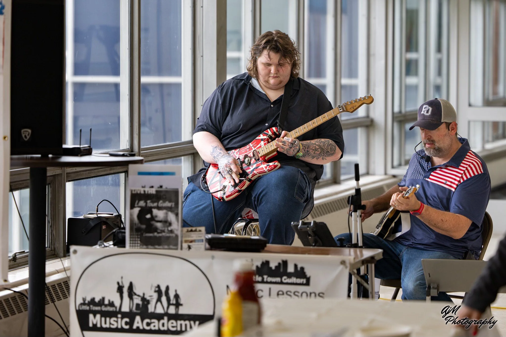 Two men playing guitars in a room with large windows, one with a decorated electric guitar and the other with an acoustic guitar, at a music event with a Little Town Guitars Music Academy banner.