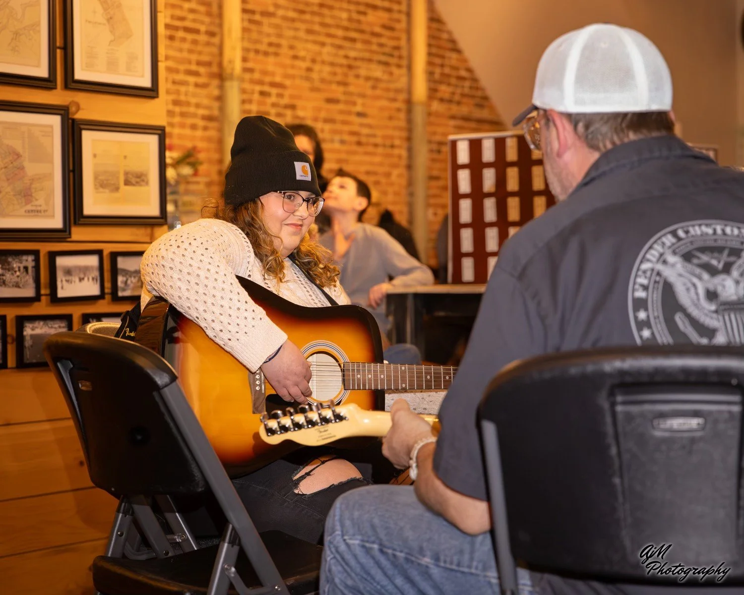 A young woman with glasses wearing a black beanie and a cream sweater, playing an acoustic guitar, sitting opposite a man with glasses and a white cap, in a cozy indoor space with brick walls and framed pictures, during a musical session.