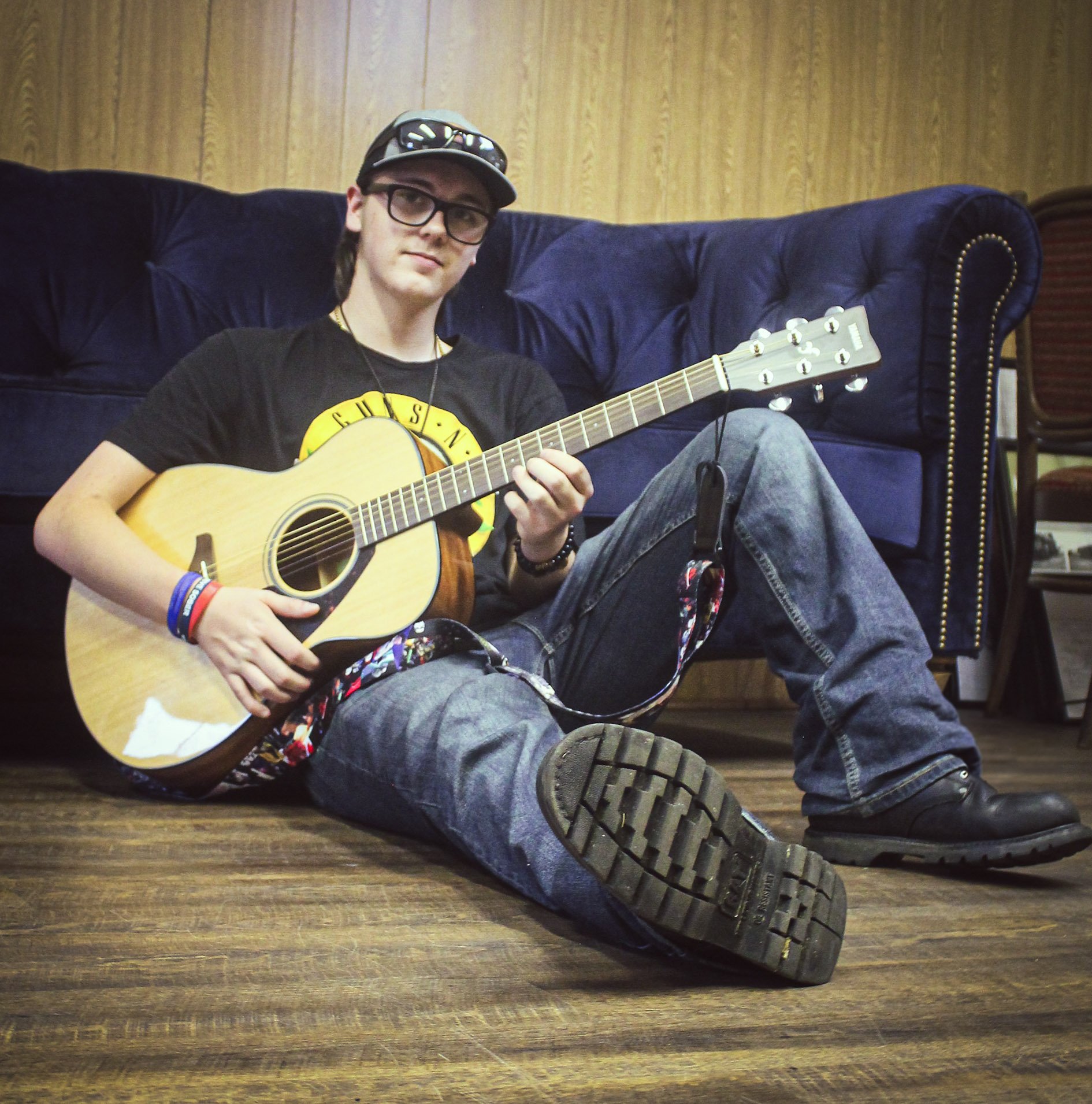 A young person sitting on the floor, playing an acoustic guitar, wearing a black Guns N' Roses t-shirt, jeans, boots, with a black cap, glasses, and wristbands.