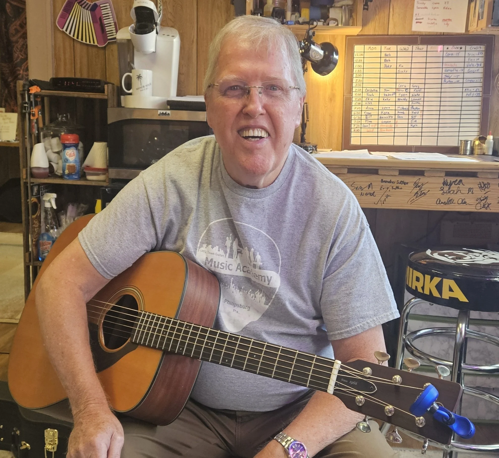 An elderly man smiling and playing an acoustic guitar in a cozy indoor setting with a warm wooden background, signed memorabilia, and a whiteboard in the background.