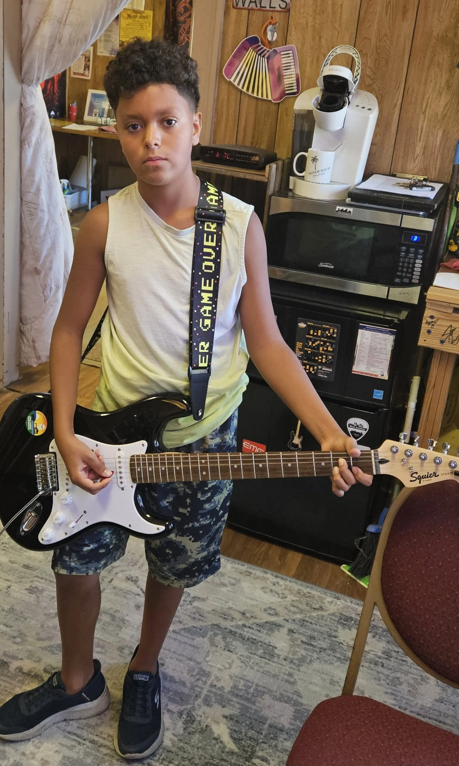 A young boy with curly hair is holding a black electric guitar in a room with wood-paneled walls. He is wearing a sleeveless shirt, camouflage shorts, and sneakers. The room contains a microwave, coffee maker, mug, and various decorations on the wall