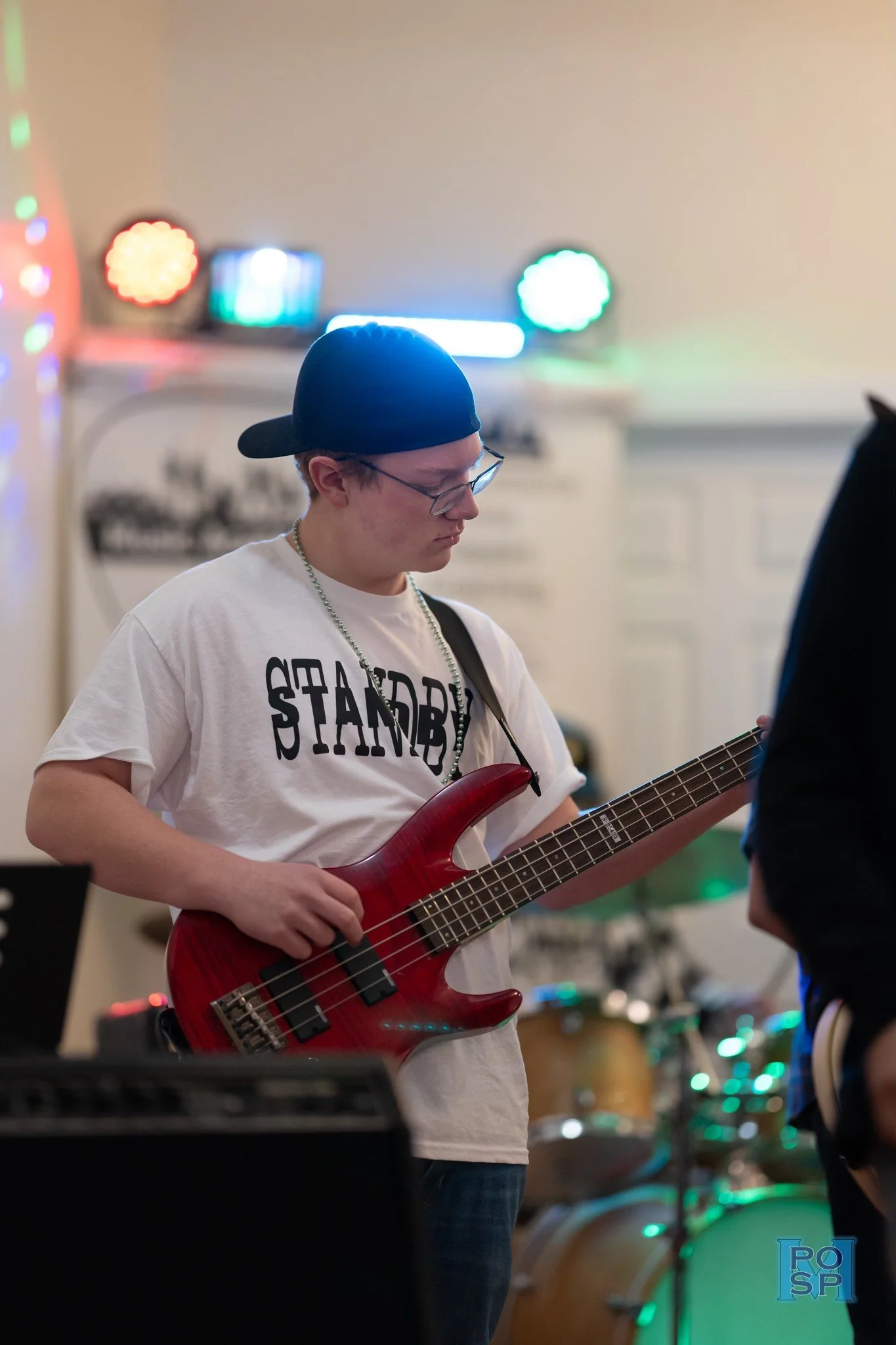 A young man with glasses and a backward cap is playing a red bass guitar at a music venue. The background shows colorful stage lights and musical instruments.