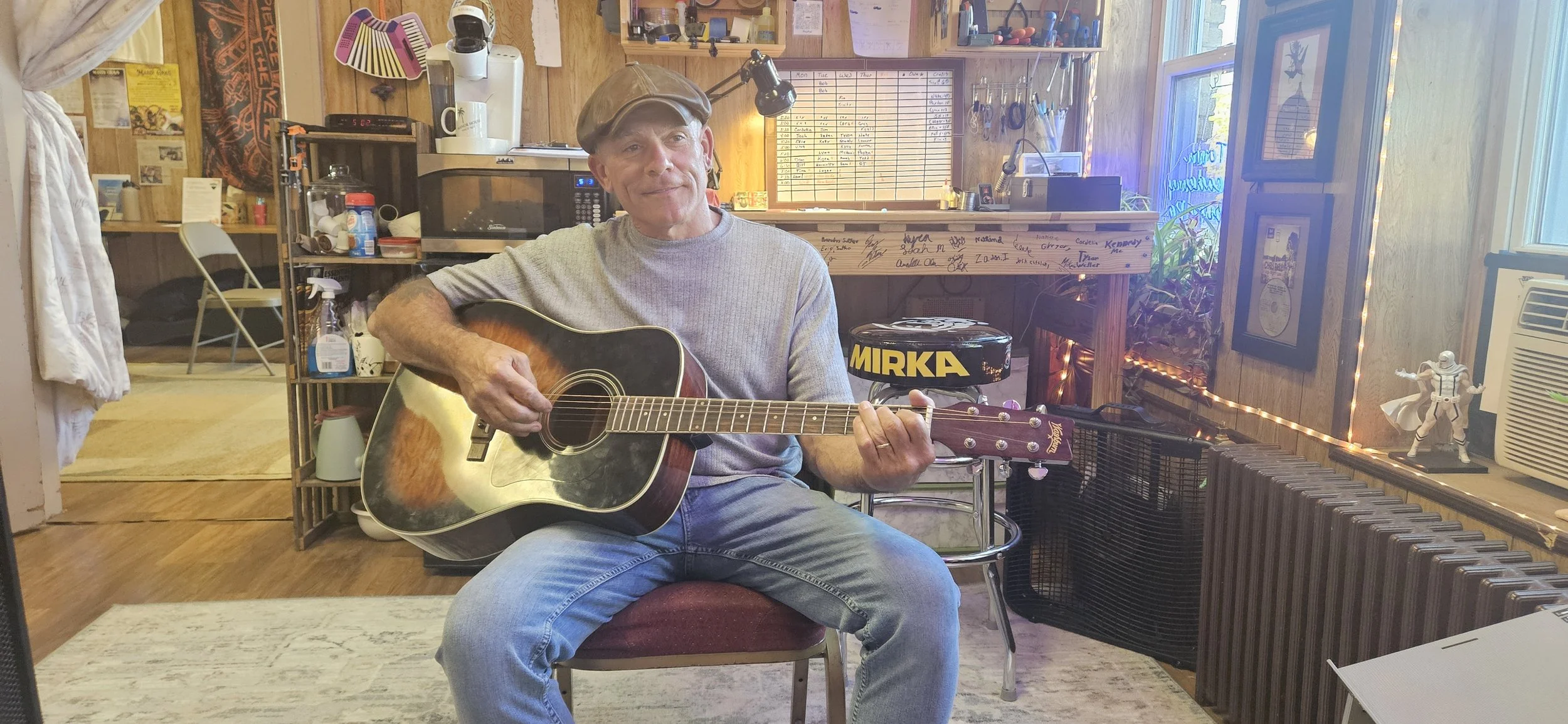 A man sitting on a red chair playing an acoustic guitar in a cozy, decorated room with wooden walls and string lights. He wears a gray t-shirt, jeans, and a brown cap, with a smile on his face.
