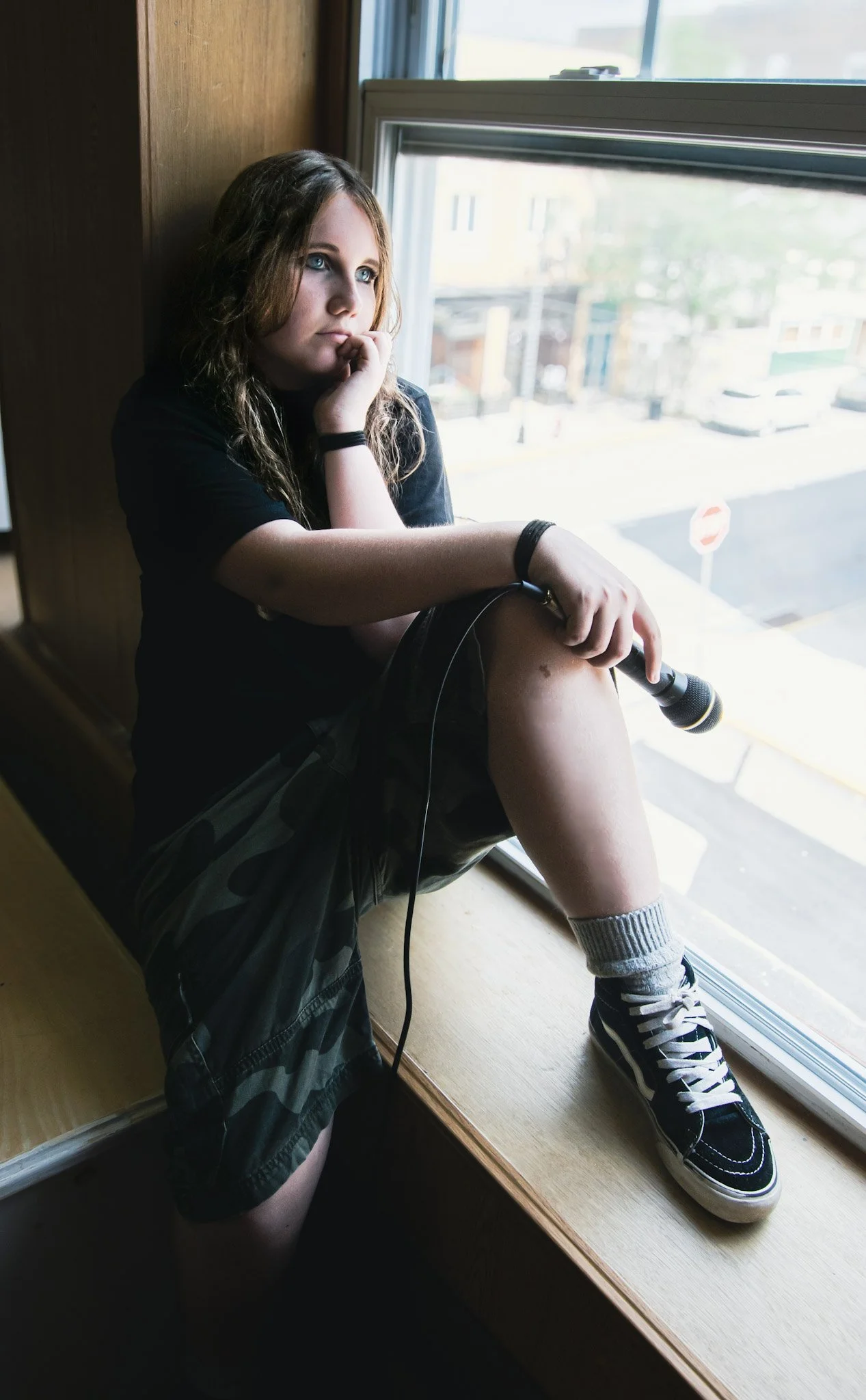 Teen girl sitting on a windowsill holding a microphone, looking out the window pensively.