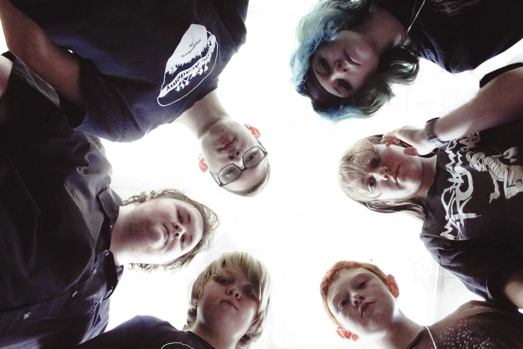 Group of seven teenagers standing in a circle and looking down at the camera, which is positioned below them, against a bright sky background.