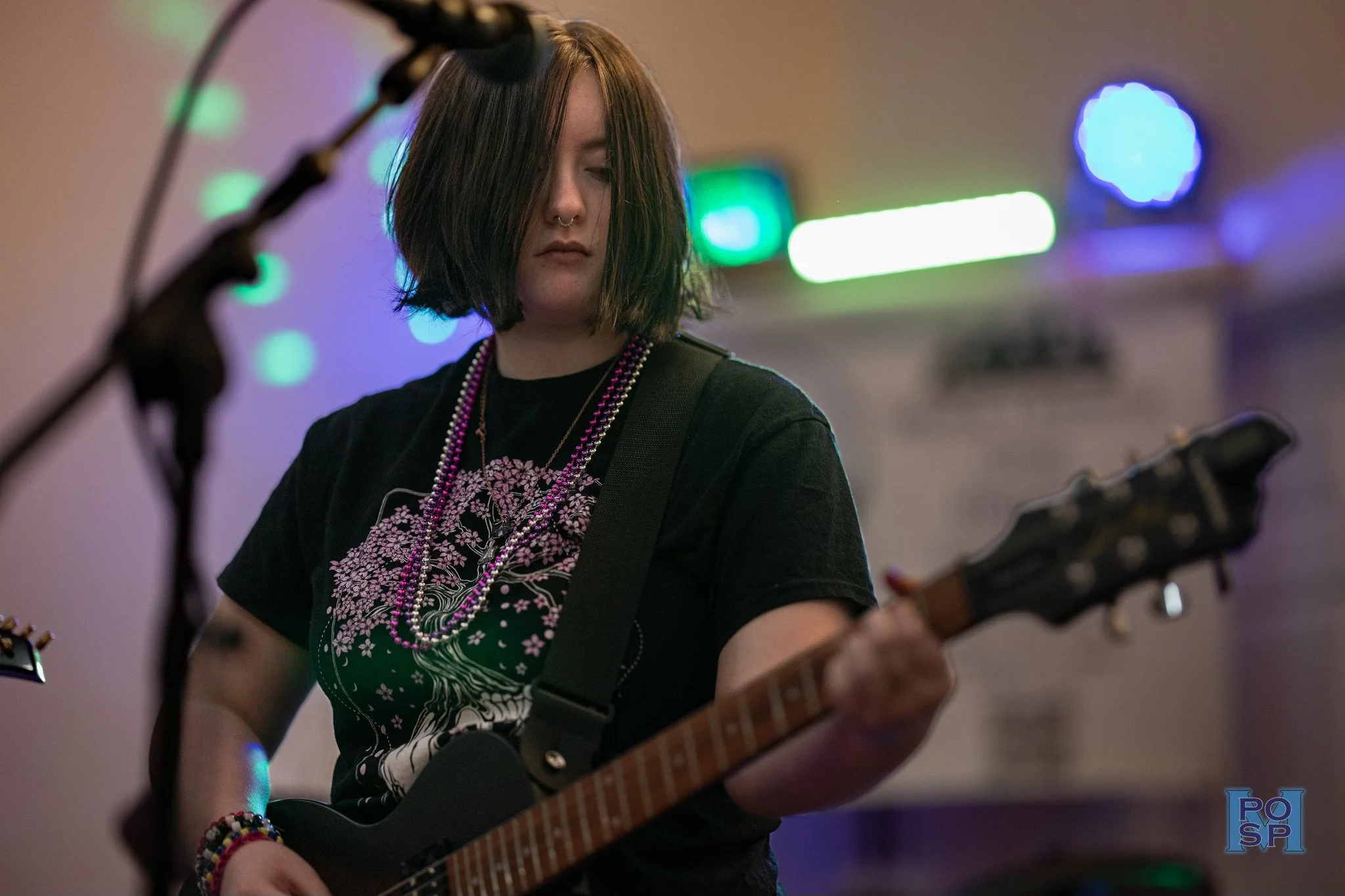 Young woman with black hair, playing electric guitar in a dimly lit room with colorful musical lights, wearing a black T-shirt with pink floral design, layered necklaces, and beads.