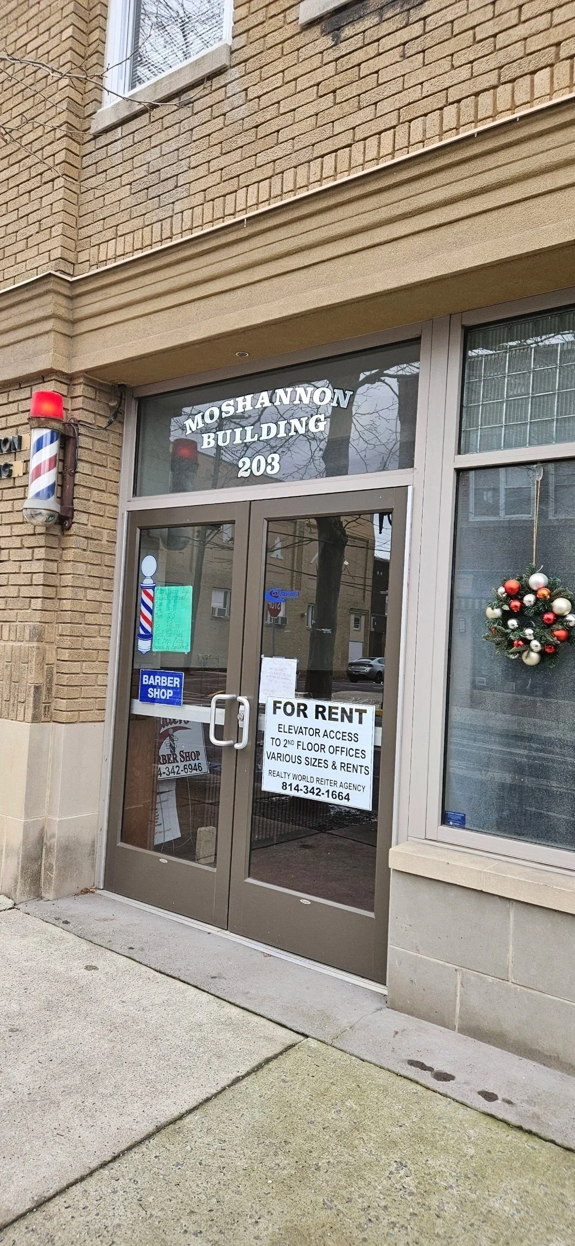 Front entrance of a building with a sign that reads 'Moshannon Building, 203.' The door has a 'For Rent' sign offering elevator access to second-floor offices, various sizes, and rents, with a contact number. There is a Christmas wreath on the window and a barber shop sign on the door. A barber pole is mounted on the wall next to the door, and the building has brown brick walls.
