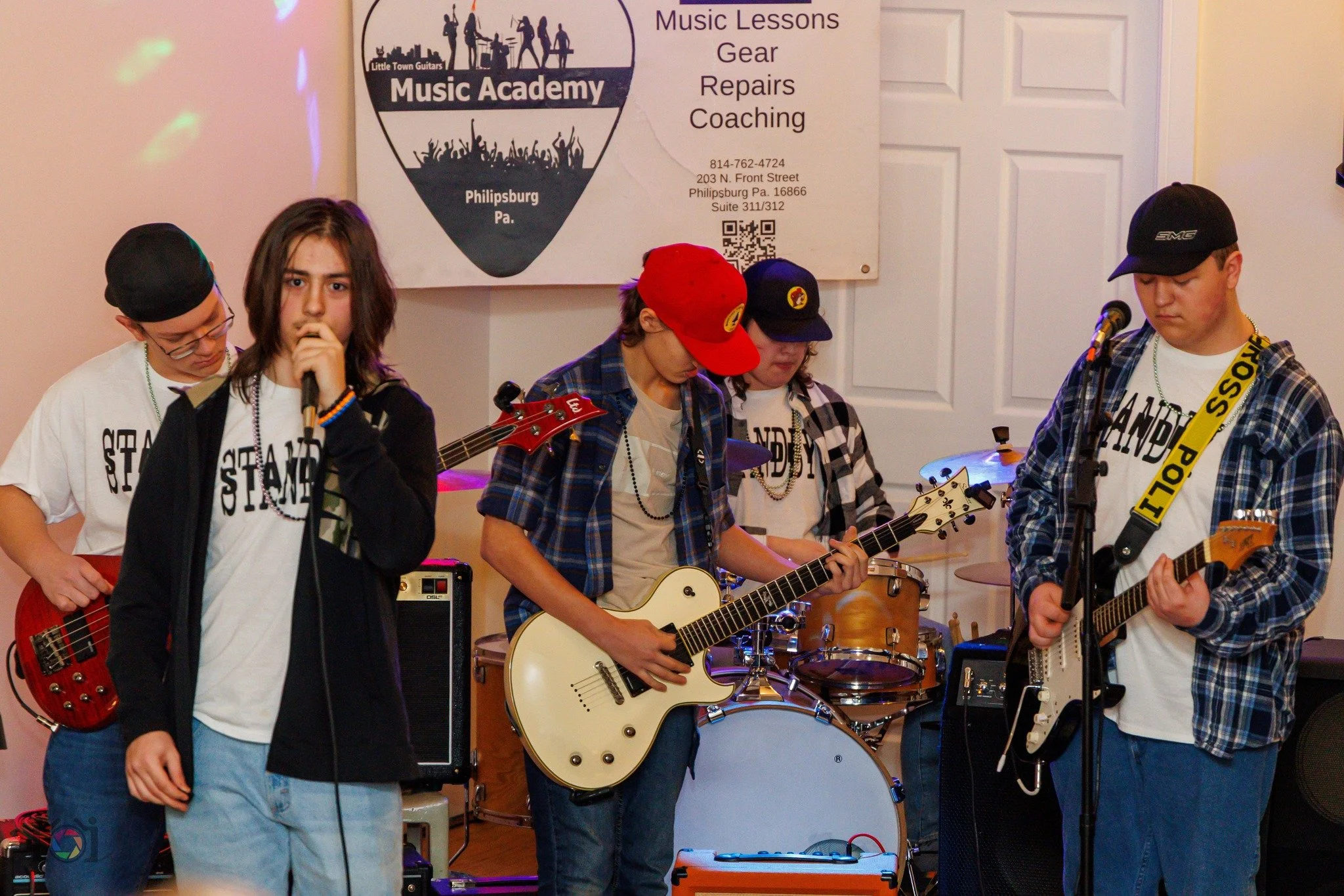 A group of young teenagers performing with musical instruments on stage, including a girl singing into a microphone, two boys playing electric guitars, and a boy on drums, with a banner advertising a music academy in the background.