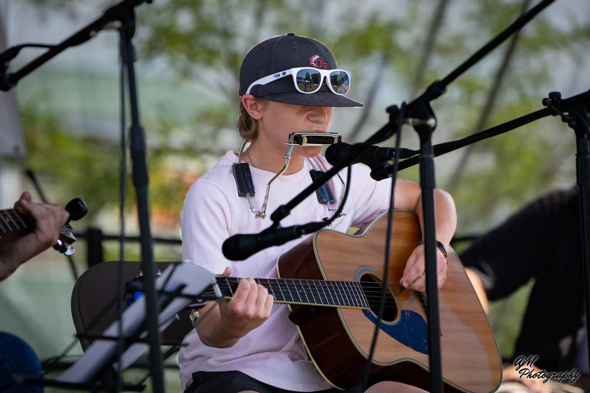 Person wearing a black cap with a red logo, white sunglasses, and a white t-shirt, playing an acoustic guitar at an outdoor event, with microphones and other musicians around.
