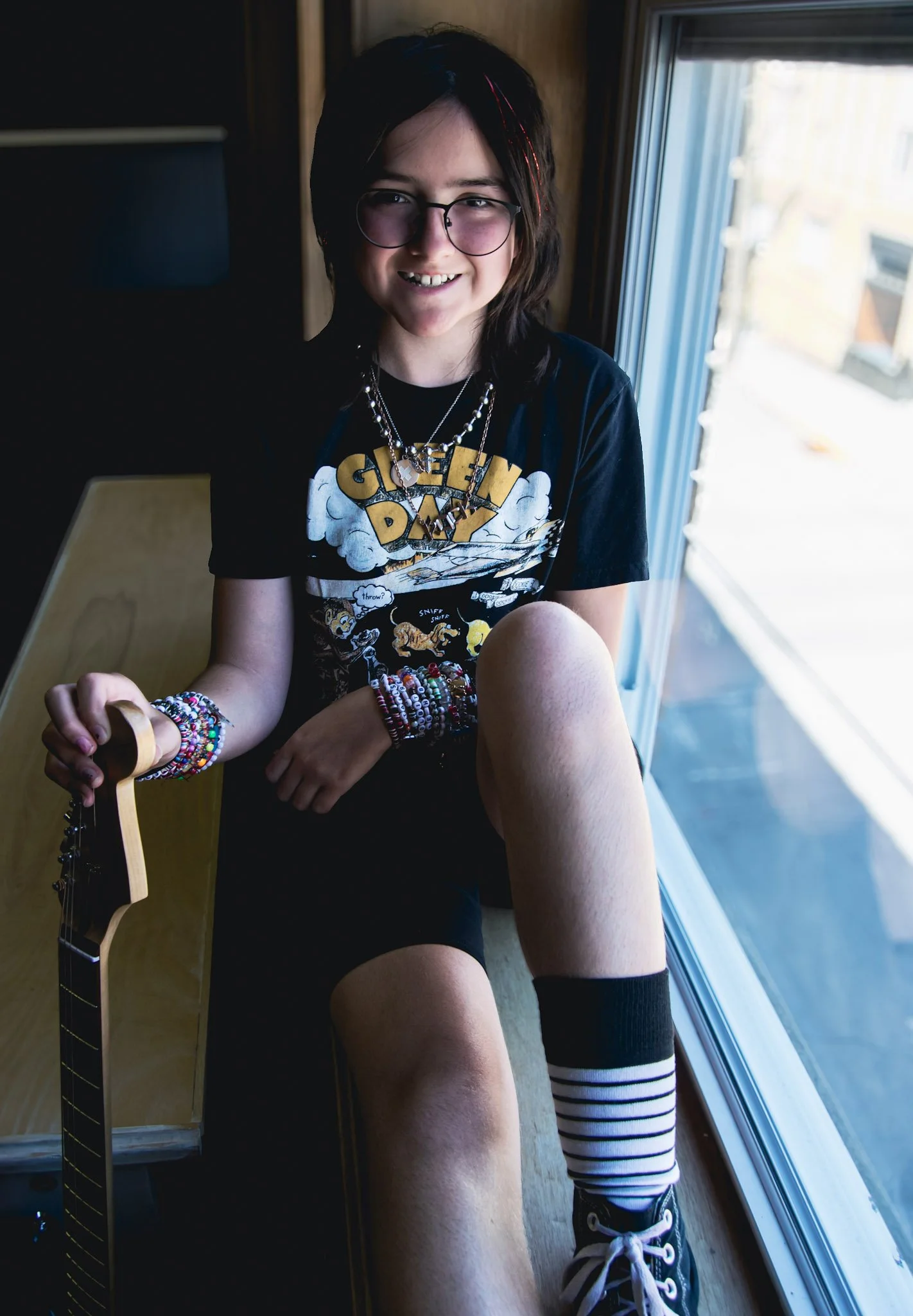 A smiling young girl with glasses and dark hair, wearing a "GEEKY DAY" T-shirt, multiple colorful bracelets, and layered necklaces, sitting on a windowsill with a guitar.