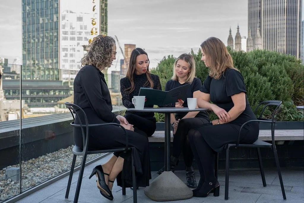 Four women sit around a small round table on a rooftop terrace with a city skyline in the background, looking at a file together, with coffee mugs on the table.