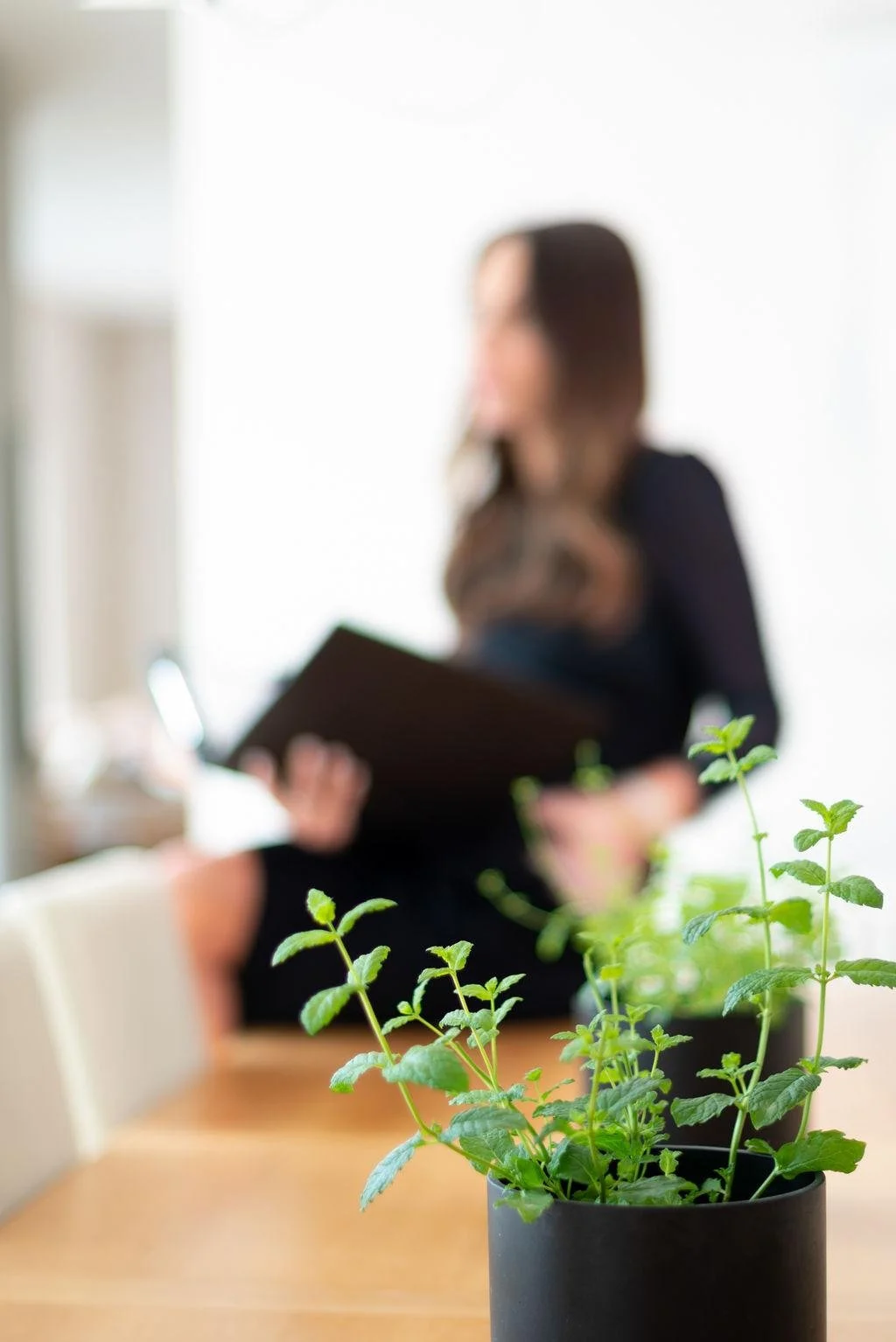 Close-up of a potted mint plant on a wooden table with blurred woman in black dress holding a notebook in background