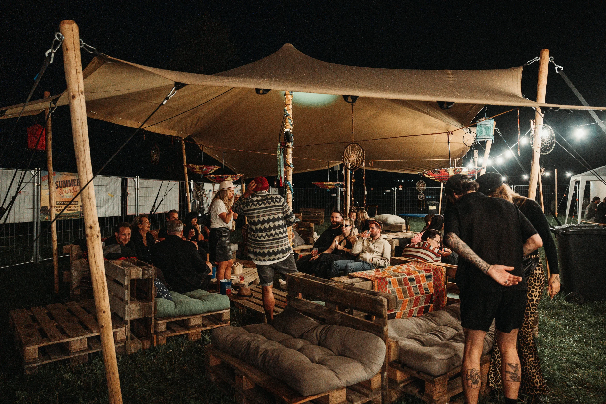 Menschen beim Treffen im Freien unter einem großen Zelt bei Nacht, mit Sitzgelegenheiten aus Paletten und Lichterketten.