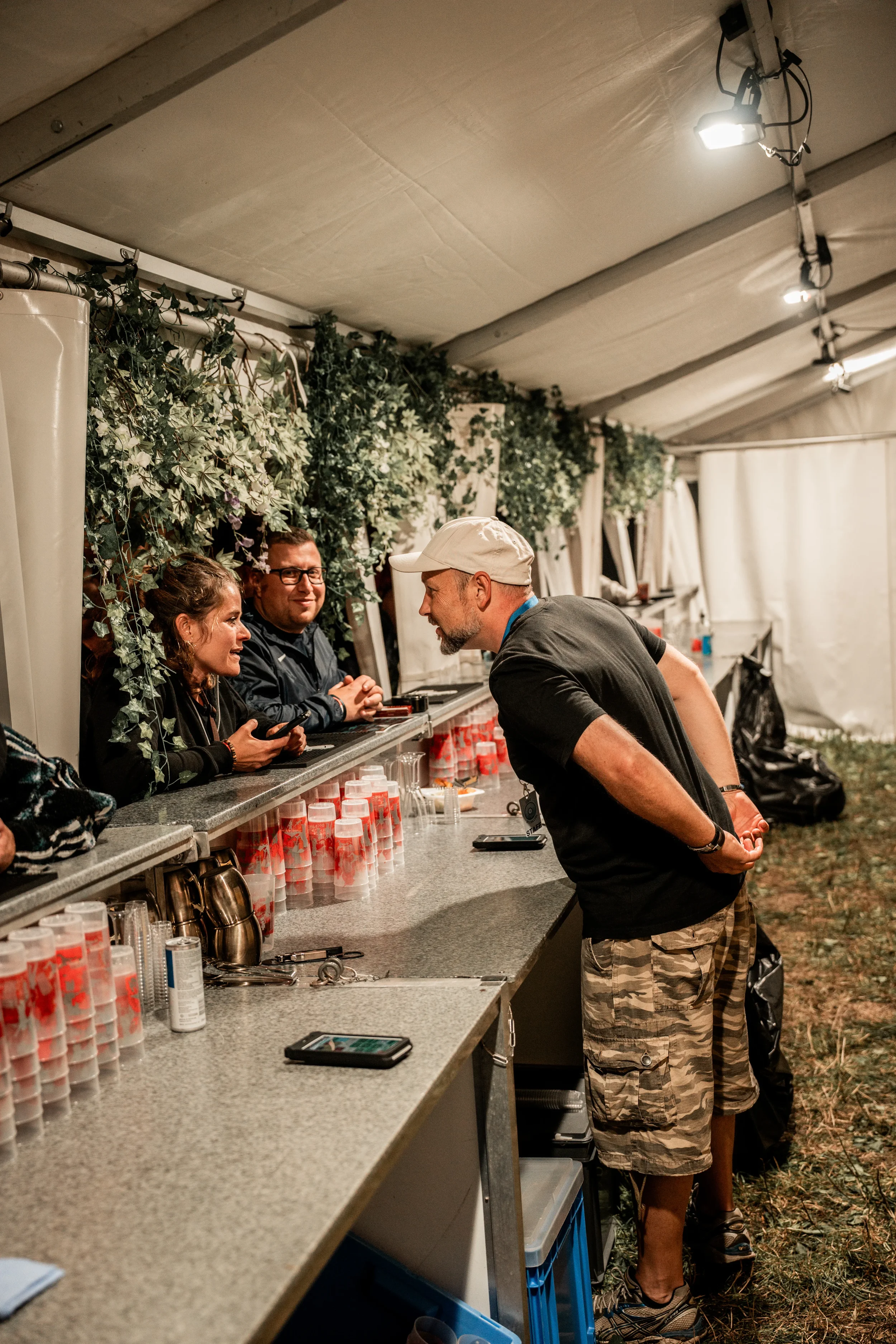 Ein Mann im Schwarzshirt und Camouflage-Hosen spricht mit zwei Frauen hinter einer Bar im Zelt bei Nacht, umgeben von grünen Pflanzen und Getränkeflaschen.