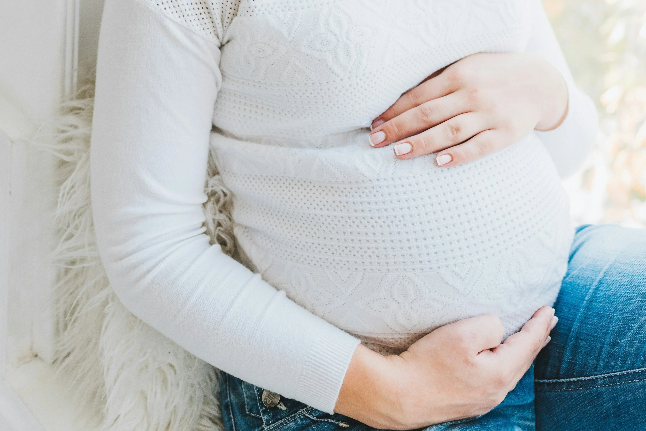 A pregnant woman wearing a white textured sweater, sitting on a white fluffy surface, with her hands gently resting on her belly.