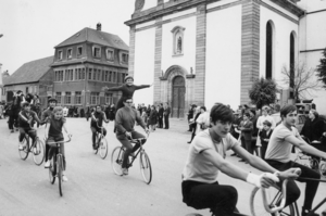Groupe d'enfants et d'adultes faisant du vélo dans une ville ancienne, avec des bâtiments historiques en arrière-plan, en noir et blanc.
