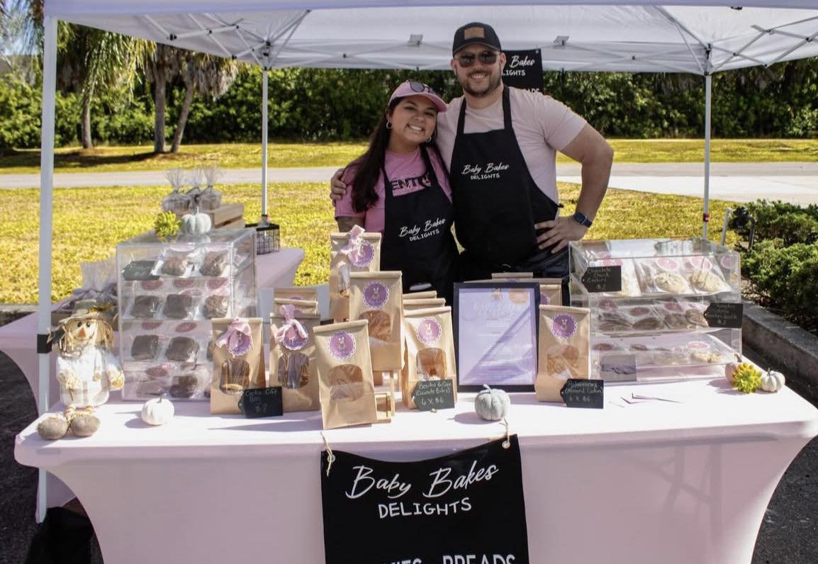Two smiling people standing behind a table at an outdoor market stall selling baked goods. They are under a white canopy, with a sign on the front of the table that reads "Baby Bakes DELIGHTS." The table displays various packaged baked items, small white pumpkins, and decorative items.