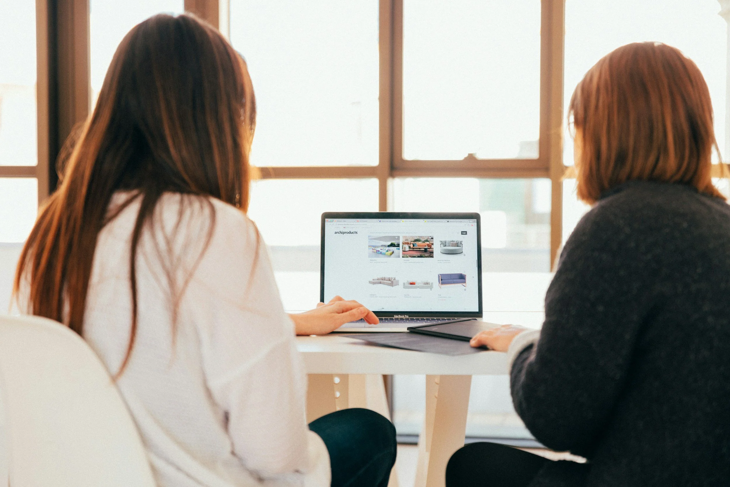 Two women sitting at a white table in front of a large window, looking at a laptop displaying furniture products.