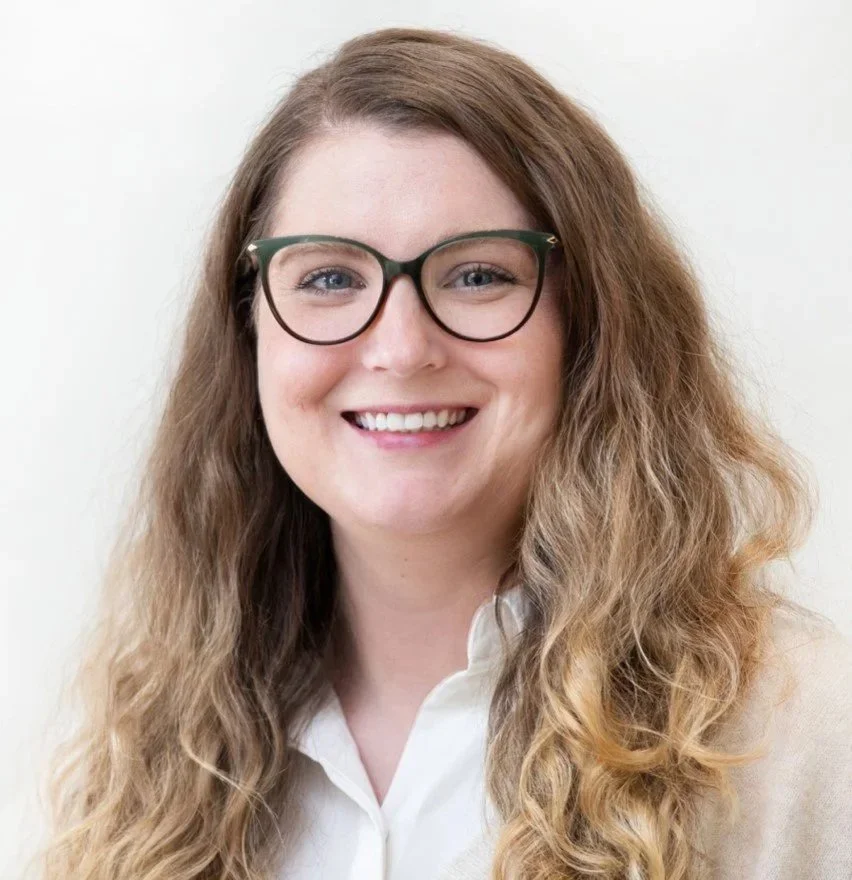 Close-up of a smiling woman with long, wavy brown hair wearing glasses and a white collared shirt.