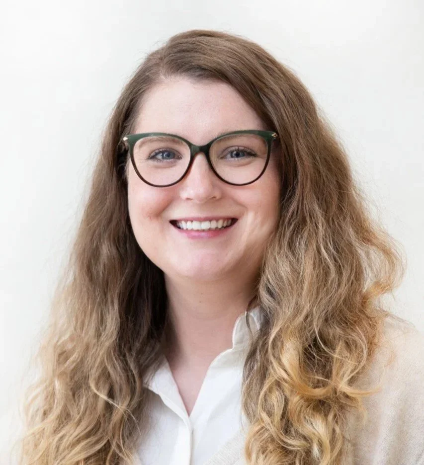 A woman with wavy, light brown hair and glasses smiling at the camera.