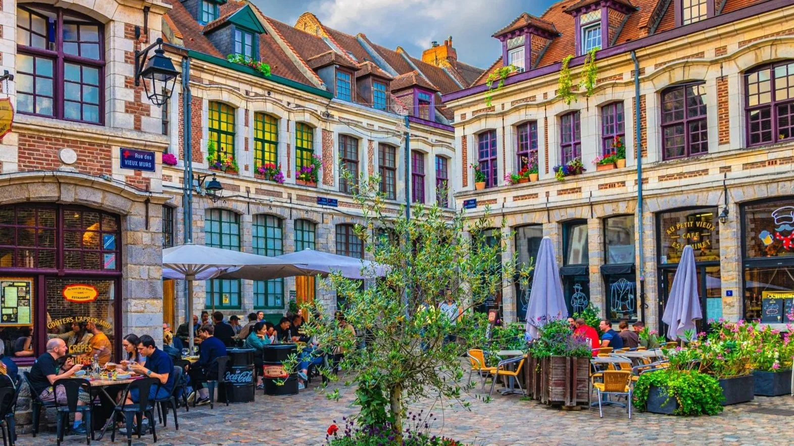 Une place pavée avec des cafés en extérieur et des bâtiments colorés de style européen autour, avec des clients assis à des tables, des parasols et des fleurs décoratives.