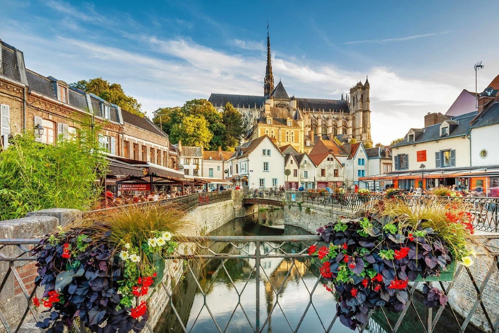 Scène urbaine de jour à Rouen avec une rivière, des maisons colorées, un pont, un bâtiment religieux gothique en arrière-plan, et des fleurs en premier plan.